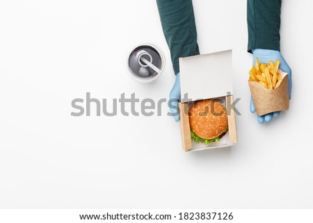 Order in fast food restaurant. Waiter in rubber gloves gives burger in cardboard box, and french fries in paper bag, glass with cola, ice and straw isolated on white background, top view, copy space
