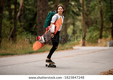 active girl riding skateboard down the road with wakeboard in her hand and backpack on her shoulders.