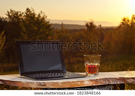 Glass with drink and laptop on a background of greenery and nature. Whiskey on an old wooden board. Sunset in summer. The concept of remote work and freelance. Rest and work in the summer.