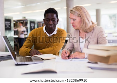 Friendly female tutor helping to diligent african-american man preparing for exam in library. High quality photo