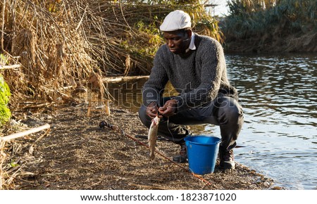Adult African fisherman putting caught fish in bucket near river