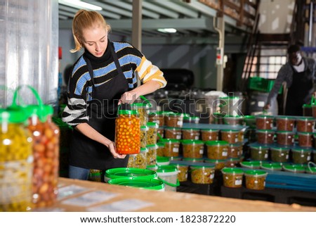 Female worker stocks plastic containers and cans with olives in warehouse. High quality photo