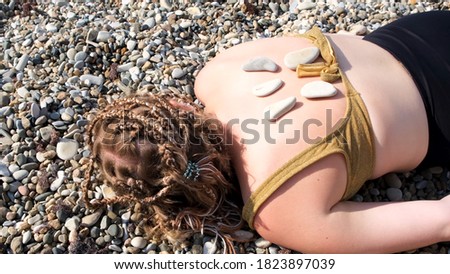 A young woman is lying on a pebble beach. Concept. Man hand putting hot stones on the back of a young girl in swimming suit with afrobraids, summer relax.