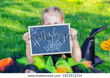 girl in a witch costume for the Halloween holiday. Placard with the inscription: Halloween. Happy girl sits on the grass and closes her eyes with a sign