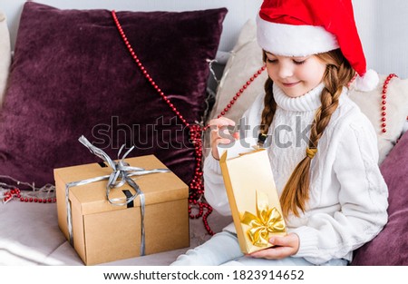 A girl in a santa claus hat sits on the sofa and opens a new year gift in a golden package