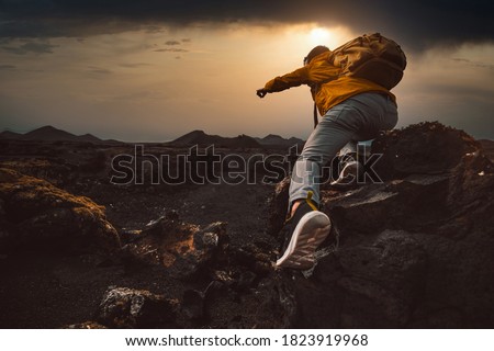 Successful hiker hiking a mountain pointing to the sunset. Wild man with backpack climbing a rock over the storm. Success, wanderlust and sport concept. Royalty-Free Stock Photo #1823919968