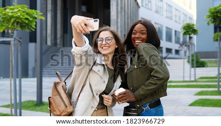 Mixed-races young pretty females smiling cheerfully to camera and taking selfie photo with mobile phone camera at city street. Multi ethnic beautiful happy women students making photos with smartphone