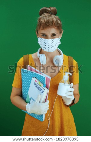 Life during coronavirus pandemic. modern learner woman in yellow shirt with disinfectant, textbooks, notebooks, medical mask, white headphones and backpack on green background.
