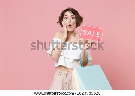 Shocked young woman 20s in summer clothes isolated on pink background. Shopping discount sale concept. Mock up copy space. Hold package bag with purchases, sign with SALE title, put hand on cheek