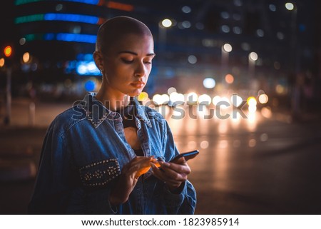 Image of a young woman using mobile phone on the street. Girl with short shaved hair walking in the evening in the city center