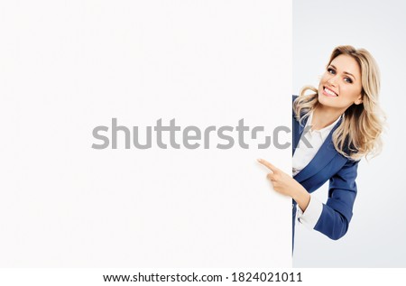 Happy smiling young businesswoman in blue confident suit, showing blank signboard with copyspace empty area for slogan or advertise text, isolated over grey background. Blond model in business concept