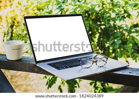 Laptop computer, glasses and coffee cup outdoor on the garden terrace. Blank screen as copy space. Greenery in the background.