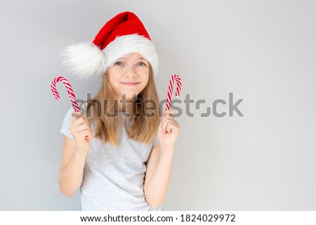 Portrait of a blonde caucasian smiling girl in a Santa hat holding two New Year's candies in her hands on a grey background. Christmas and happines concept. Close up, copy space