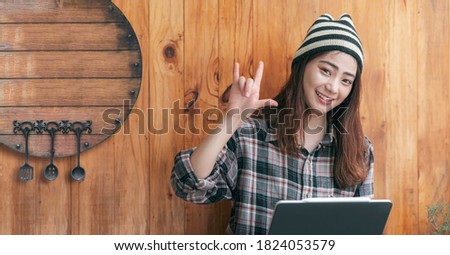 Portrait of young female teenager wearing wool hat and plaid shirt showing i love you hand sing, smiling and looking at camera while standing near wooden wall, copy space.
