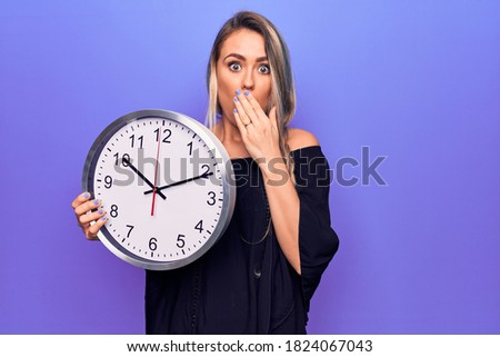 Young beautiful blonde woman doing countdown using big clock over purple background covering mouth with hand, shocked and afraid for mistake. Surprised expression