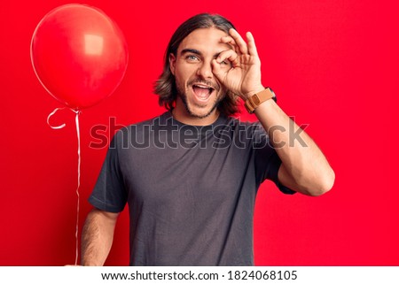 Young handsome man holding balloon smiling happy doing ok sign with hand on eye looking through fingers 