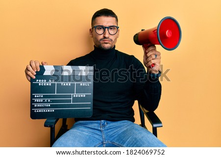 Handsome man with tattoos holding video film clapboard and megaphone skeptic and nervous, frowning upset because of problem. negative person.  Royalty-Free Stock Photo #1824069752