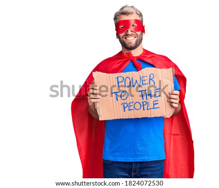 Young blond man wearing super hero custome holding power to the people cardboard banner looking positive and happy standing and smiling with a confident smile showing teeth 