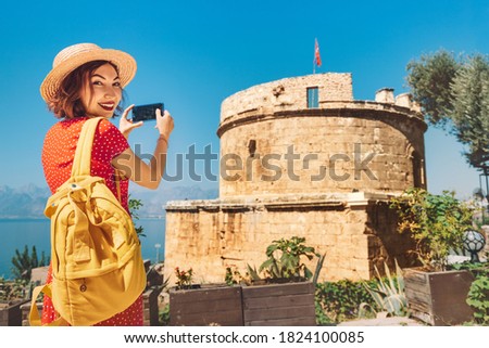 Happy woman traveler takes photos on her mobile phone of the sights of the Mediterranean part of Turkey. Ancient fortress tower and the sea in the background