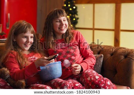 two teenage girls with loose hair in red pajamas are watching a movie, laughing and eating popcorn.
