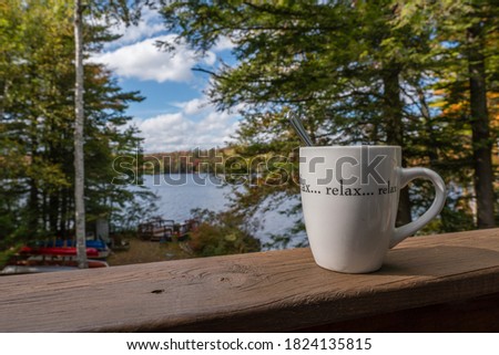 Close-up of mug with beverage on railing with the word "relax" on it with a lake and beautiful fall foliage in background