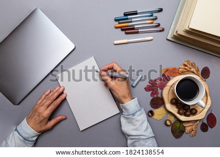 Cozy home office concept. Hands of an elderly woman with a pen about to write something in a notebook. Laptop, books, pens and coffee cup with autumn leaves top view with copy space. 