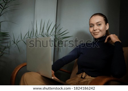 young woman of Caucasian appearance works reads an article writes plays on a laptop computer, sits in a comfortable chair, modern clothes for the office. Thinks about a successful career in business.
