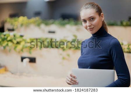 looking at the camera, young confident woman holding a laptop computer in her hands, completing a new business task. stylish office clothes, hair pulled back, blonde Caucasian appearance.