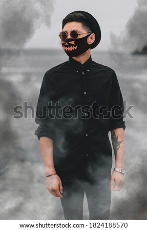 Stylish young Asian man in a black medical mask with a picture of a pumpkin, style for the celebration of Halloween 2020 during the coronavirus pandemic. Quarantine, stylish image, smoke, mysticism