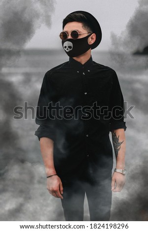 Stylish young Asian man in a black medical mask with a picture of a skull, style for the celebration of Halloween 2020 during the coronavirus pandemic. Quarantine, stylish image, smoke, mysticism