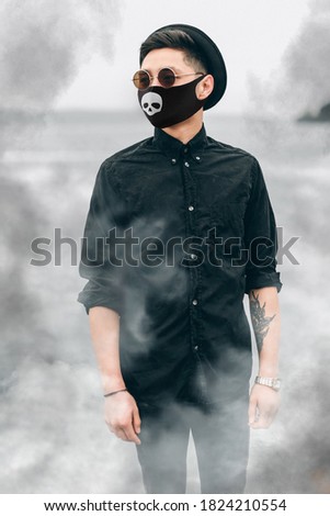 Stylish young Asian man in a black medical mask with a picture of a skull, style for the celebration of Halloween 2020 during the coronavirus pandemic. Quarantine, stylish image, smoke, mysticism
