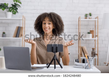 Modern blogger work and hobby during covid-19 epidemic. Cheerful african american female records video for followers and looks at webcam of smartphone at table with laptop in interior of living room