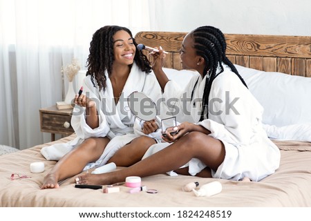 Joyful black women in bathrobes sitting on bed at home, putting makeup, looking at mirror. Attractive african american female friends having fun together at home, trying beauty products, copy space Royalty-Free Stock Photo #1824248519
