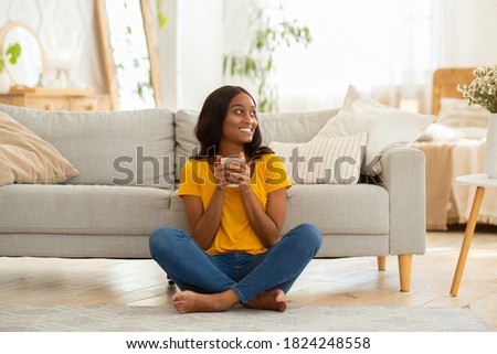 Full length portrait of young black woman drinking coffee on floor near sofa at home. Cheerful African American lady enjoying aromatic hot beverage in living room