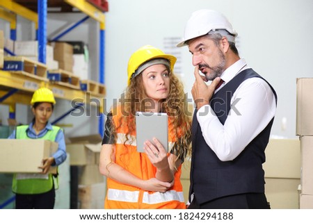 Caucasian male boss with his female store manager in full safety equipment are checking for the inventory using digital tablet inside warehouse while the employee working in background