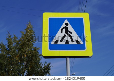 A pedestrian crossing sign against a blue sky and a yellow-green tree. Close-up.