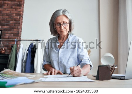 Smiling middle aged stylish woman fashion designer drawing sketches in studio. Mature old adult elegant grey-haired lady dressmaker small business owner creating new fashion design cloth in atelier.