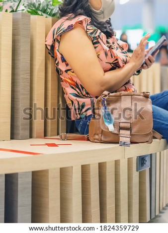 Mini alcohol gel bottle to kill Corona Virus(Covid-19) hang on a leather shoulder bag of a woman wearing a mask while use smartphone with alternative seating mark for social distancing in the mall.
