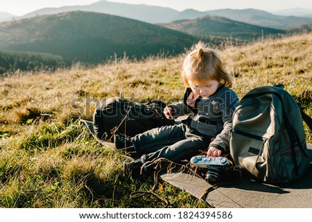 Little hiker girl with camera and backpack sitting on grass in autumn mountains. Travel concept. Holiday, trip and vacation. World Tourism Day.