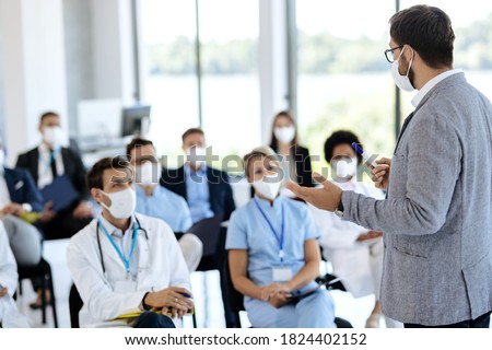 Male entrepreneur holding a training class to large group of healthcare workers and business people in conference hall. They are all wearing protective face mask due to COVID-19 pandemic.  Royalty-Free Stock Photo #1824402152