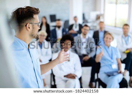 Young doctor talking to group of his colleagues and business people during presentation in convention center.  Royalty-Free Stock Photo #1824402158