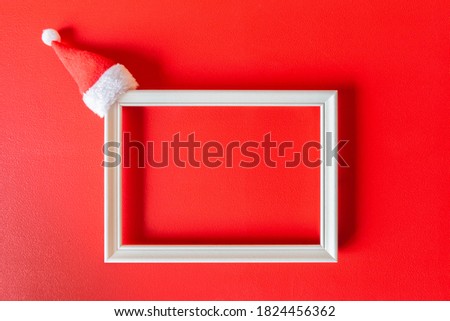White frame with small Santa Claus hat on red painted background. Minimalist festive decoration with copy space. Mockup for Christmas time. Christmas, New Year, winter concept.