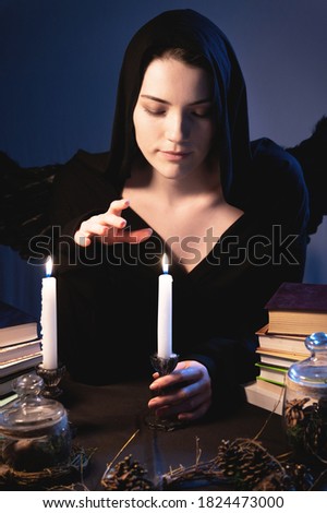 Medium closeup attractive Caucasian girl with golden long hair in a black hood sits at a table and leafs through a book. Costume with wings. Candles are burning. Halloween celebration concept