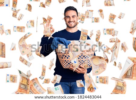 Young handsome man holding laundry basket doing ok sign with fingers, smiling friendly gesturing excellent symbol