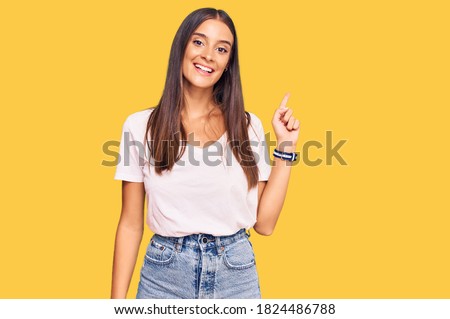 Young hispanic woman wearing casual white tshirt with a big smile on face, pointing with hand finger to the side looking at the camera. 