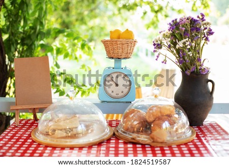 Pastries and pies on window of a bakery-coffee shop with beautiful old scales. Showcase of an street summer cafe with pastries. Croissants and buns on checkered tablecloth on terrace. Veranda decor