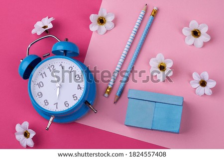 Stationery accessories on the pink background. Alarm clock, pencils and blue container for chalks on the table.