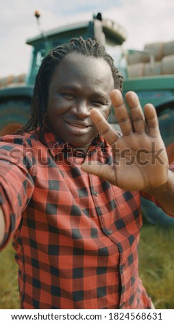 Video call. African farmer waving to the smartphone camere in front of the tractor. High quality photo