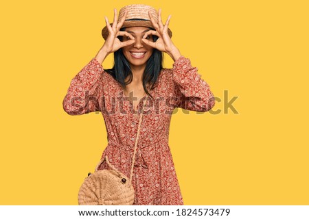 Young african american woman wearing summer hat doing ok gesture like binoculars sticking tongue out, eyes looking through fingers. crazy expression. 