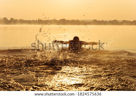 Sporty young man actively swimming at lake and making splashes on water. Silhouette of strong guy doing exercises at morning time on fresh air.
